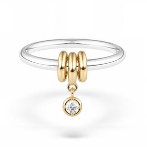 May include: A silver-toned ring band featuring three small, smooth gold-toned rings threaded onto it. A single, circular gold-toned charm set with a clear, faceted gemstone hangs from the center ring, creating a delicate and polished appearance.