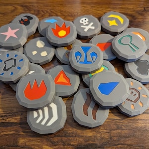 May include: A collection of gray, circular game tokens with various colorful symbols. The tokens feature designs such as flames, stars, and geometric shapes in red, orange, yellow, blue, and white. The tokens are arranged on a wooden surface.