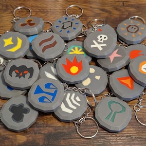 May include: A collection of gray, stone-shaped keychains with various colorful geometric designs. Each keychain has a silver keyring attached. The designs include a skull, flames, and other abstract shapes. The keychains are arranged on a wooden surface.