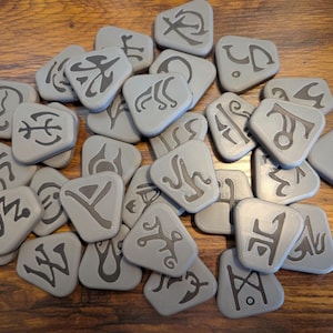 May include: A collection of gray, triangular rune stones with dark gray symbols. The stones are scattered on a wooden surface, showcasing a variety of ancient-looking characters. The runes are likely used for divination or spiritual practices.