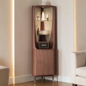 May include: A corner display cabinet made of wood with a glass front. The cabinet is a warm brown color and has a fluted design on the lower section. Inside, there are shelves holding decorative items, including vases and a speaker.