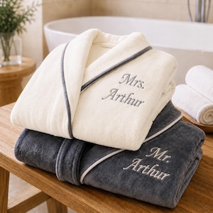 Puede incluir: Una bata de felpa color crema con ribetes grises y el texto bordado Mrs. Arthur, sobre una bata de felpa gris oscuro con ribetes crema y el texto bordado Mr. Arthur, ambas con mangas largas y un diseño de cuello tipo chal.