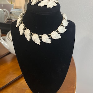 May include: A jewelry set consisting of a necklace and matching clip-on earrings. The necklace features a series of off-white, carved leaf-shaped links connected by gold-tone metal hinges. The earrings mirror the leaf design in the same off-white color.