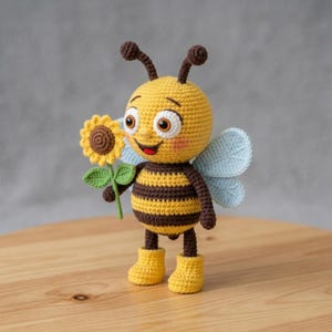 Op de afbeelding: Een gehaakte bijenpop met een gele body, bruine strepen en blauwe vleugels. De bij houdt een zonnebloem vast met een bruin hart en groene bladeren. De pop draagt gele laarzen en heeft een lachend gezicht.
