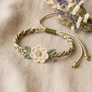 May include: A braided bracelet featuring a central cream-colored crocheted flower with a gold-tone bead center, flanked by two small green crocheted leaves. The band incorporates green and cream cord with small gold-tone beads and adjustable ends.