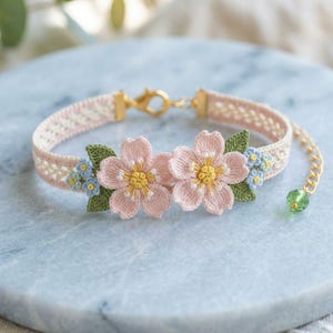 Op de afbeelding: Een delicate roze en crèmekleurige armband met twee geborduurde roze bloemen met gele centra en groene bladeren. De armband heeft een gouden sluiting en een kleine groene kralenbedel. De armband is ongeveer 15 cm lang.
