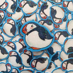 Peut inclure: Un ensemble d'autocollants représentant un macareux moine de dessin animé. Les macareux ont un corps blanc, des ailes bleu foncé et un bec orange. Chaque autocollant a une bordure bleue et une finition scintillante et irisée.