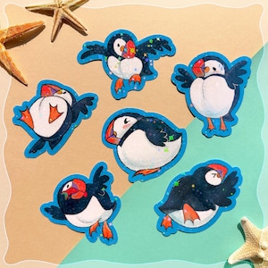 May include: A collection of six stickers featuring cartoon puffins in various poses. The puffins have black and white bodies, orange feet, and red and orange beaks. Each sticker has a blue outline with a glittery effect, and the background is a sandy color.