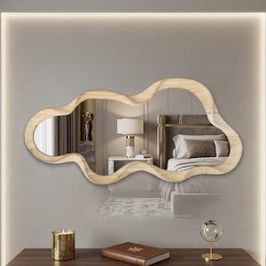 May include: A cloud-shaped mirror with a light wood frame hangs on a gray wall. The mirror reflects a bedroom scene. A gold candle, a book, and a decorative sphere sit on a dark wood table below.