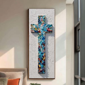 May include: A textured, vertical artwork featuring a cross. The cross is painted with a palette of blues, reds, and yellows, creating a vibrant, impressionistic effect. The background is white, and the artwork is framed in a simple, dark frame. The piece is displayed on a wall.