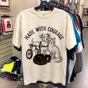 May include: Cream-colored t-shirt featuring a graphic of a dog in a wheelchair, a striped cat, and a black cat. The text "MADE WITH COURAGE" is arched above the animals. The shirt is hanging on a rack with other clothing.