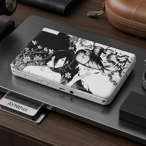 May include: A white handheld gaming console with a black and white manga-style illustration. The artwork features a character with long hair and floral elements. The console is on a metallic surface with accessories.