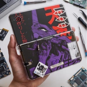 Puede incluir: Una funda transparente y articulada para consola de juegos portátil con un diseño de anime morado y negro. La funda tiene un gráfico de robot y texto japonés. También se ve un pequeño cartucho de juego blanco.