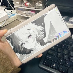 Puede incluir: Una consola de juegos portátil con un marco beige y una pantalla que muestra una ilustración de estilo anime en blanco y negro de una chica con ojos morados. La pantalla también tiene la palabra "EYES" en letras holográficas.