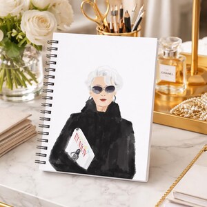 May include: A spiral-bound notebook featuring an illustration of a woman in sunglasses and a black coat, holding a magazine with the word "Runway." The notebook is on a marble surface, with a gold-toned lamp and perfume bottle in the background.