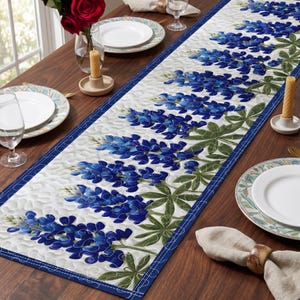 May include: A quilted table runner featuring a repeating pattern of bluebonnet flowers with green leaves on a white background. The runner has a blue border and is placed on a wooden table, alongside plates, glasses, and candles.