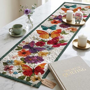 May include: A white quilted table runner with a green border, adorned with colorful butterfly and floral appliqués. The runner is decorated with orange, red, purple, and blue butterflies and flowers. A tag reads "Handcrafted & Unique".