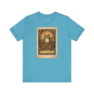 May include: A light blue t-shirt featuring a whimsical illustration of a frog holding a mushroom, labeled "THE CHAOS GREMLIN." The artwork is set within a decorative frame, creating a unique and playful design.