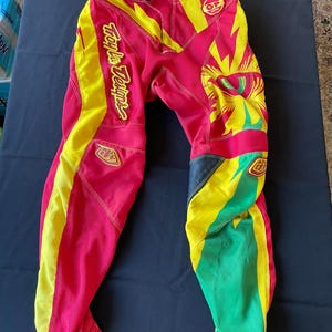 May include: Red, yellow, and green motocross pants with the words "Troy Lee Designs" in yellow script. The pants have a vibrant design with geometric shapes and accents. The pants are made of a durable material.