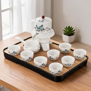 May include: A white ceramic tea set on a bamboo tray. The set includes a teapot, a serving pitcher, and eight small cups. Each cup and the teapot feature a blue and green mountain landscape design. The tray is rectangular with a black border.