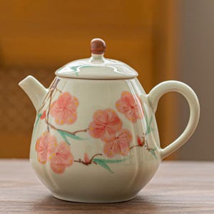 Puede incluir: Tetera de cerámica color crema con tapa y asa, decorada con flores de cerezo rosas y hojas verdes. La tapa tiene un pequeño pomo de madera marrón. Objeto decorativo.