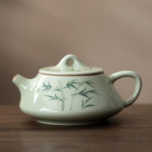 May include: A pale green ceramic teapot with a lid and handle. The teapot features a bamboo design in green. The teapot is placed on a wooden surface.