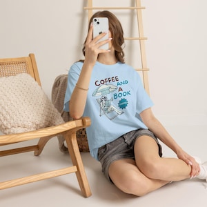 Puede incluir: Una camiseta azul claro con un estampado gráfico de una persona sentada en una silla de playa bajo una sombrilla, leyendo un libro y sosteniendo una taza. El texto dice COFFEE AND A BOOK con una estrella azul que contiene la palabra SUMMER.