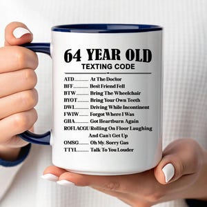 May include: White ceramic mug with a blue interior and handle, featuring the text "64 YEAR OLD TEXTING CODE" and a list of texting abbreviations and their meanings. The mug is held by a person.