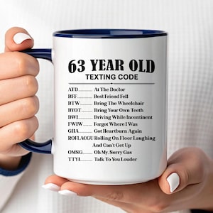 May include: White ceramic mug with a navy blue interior and handle. The mug features the text "63 YEAR OLD TEXTING CODE" in black, along with a list of texting acronyms and their meanings. A humorous gift idea.