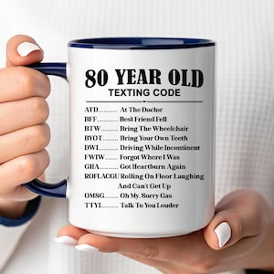 May include: White ceramic mug with a blue interior and handle, featuring the text "80 YEAR OLD TEXTING CODE" and a list of acronyms with their meanings. The mug is held by a person.