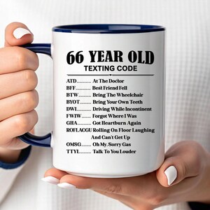 May include: White ceramic mug with a navy blue interior and handle. The mug features the text "66 YEAR OLD TEXTING CODE" in black, along with a list of acronyms and their meanings. The mug is held by a person.