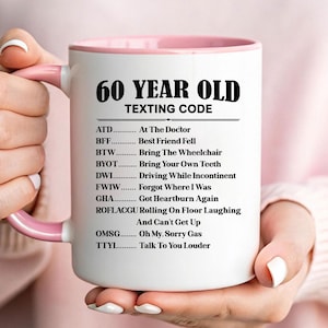 May include: A white ceramic mug with a pink interior and handle. The mug features the text "60 YEAR OLD TEXTING CODE" in black, along with a list of acronyms and their meanings, such as "ATD... At The Doctor."