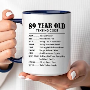 May include: White ceramic mug with a navy blue interior and handle, featuring the text "89 YEAR OLD TEXTING CODE." The mug lists humorous texting abbreviations related to aging, such as "Bring The Wheelchair" and "Forgot Where I Was."