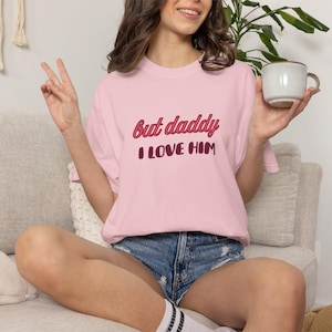 Könnte beinhalten: Ein hellrosa kurzärmeliges T-Shirt mit dem Text "but daddy I LOVE HIM", der in roter und dunkelroter Schrift auf der Brust gedruckt ist. Das Shirt hat einen Rundhalsausschnitt und eine lockere Passform aus einfarbigem Baumwollstoff.