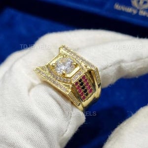 以下が含まれることがあります： 中央に大きな正方形の透明な石が付いたゴールドカラーのリング。リングには、より小さな透明な石と赤い石がちりばめられています。青い背景に、手袋をはめた手がリングを持っています。「TDJEWELS」のブランド名が表示されています。