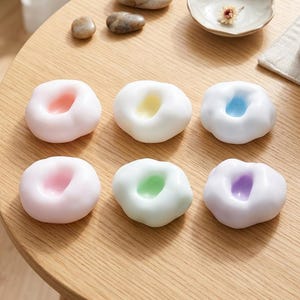 Peut inclure: Six petits objets blancs en forme de nuage avec des centres aux couleurs pastel : rose, jaune, bleu, rose, vert et violet. Les objets sont disposés sur une surface en bois brun clair, à côté de petites pierres et d'une petite coupelle blanche.