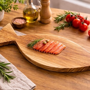 May include: A wooden fish-shaped serving board with sliced salmon, tomatoes, and rosemary. The board is on a wooden table, with a small bowl of pink salt, olive oil, and a potted plant in the background.