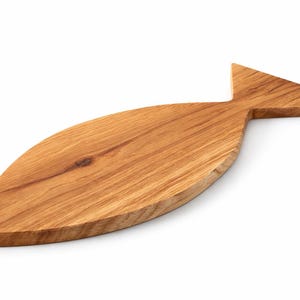 May include: A wooden cutting board in the shape of a fish. The board is made of light brown wood with visible grain patterns. The fish-shaped board has a tail fin and a rounded body, perfect for serving appetizers or preparing food.