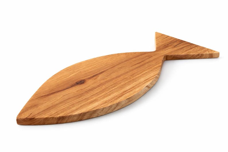 May include: A wooden cutting board in the shape of a fish. The board is made of light brown wood with visible grain patterns. The fish-shaped board has a tail fin and a rounded body, perfect for serving appetizers or preparing food.