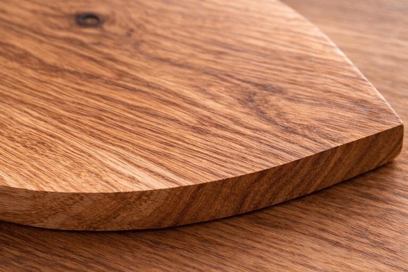 May include: Close-up of a wooden cutting board with a curved edge. The board is made of light brown wood with visible grain patterns. The surface is smooth and appears to be well-finished, suggesting a quality kitchen item.