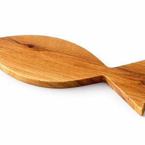 May include: A wooden cutting board in the shape of a fish. The board is made of light brown wood with visible grain and a natural finish. The fish-shaped board has a rounded body and a triangular tail.