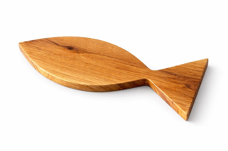 May include: A wooden cutting board in the shape of a fish. The board is made of light brown wood with visible grain and a natural finish. The fish-shaped board has a rounded body and a triangular tail.
