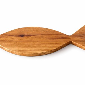 May include: A wooden cutting board in the shape of a fish. The board is made of light brown wood with visible grain and a natural finish. The fish-shaped board has a rounded body and a triangular tail.