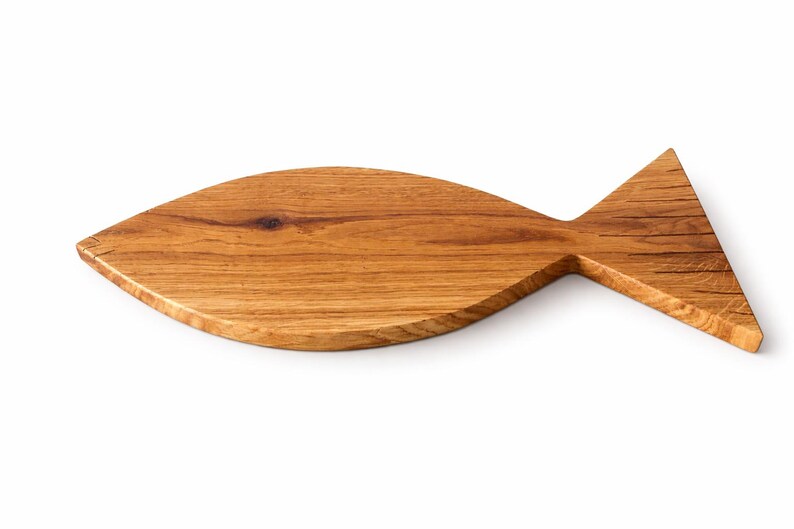 May include: A wooden cutting board in the shape of a fish. The board is made of light brown wood with visible grain and a natural finish. The fish-shaped board has a rounded body and a triangular tail.