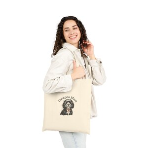 May include: Cream-colored tote bag featuring a watercolor illustration of a Cavapoo dog with the text "Cavapoo Mum". The dog has black and white fur and a red collar. The bag is held by a person.