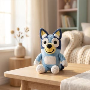 Può includere: Un peluche di Bluey all'uncinetto fatto a mano, con un corpo blu e azzurro e dettagli bianchi. Il giocattolo ha grandi occhi, una bocca gialla sorridente ed è seduto su un tavolo di legno. Lo sfondo include un vaso con fiori secchi.
