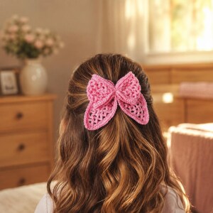 以下が含まれることがあります： 明るいピンク色のかぎ針編みのヘアアクセサリーで、質感のあるオープンワークパターンのリボン型をしています。リボンは長くウェーブのかかった茶色の髪の中央に留められており、中央の結び目の両側に2つのループがあるレイヤードデザインが特徴です。