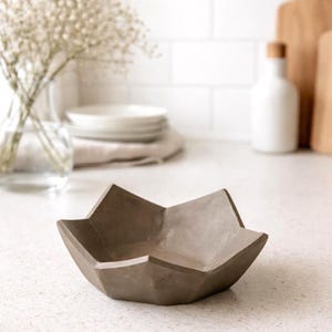 Könnte beinhalten: Eine sternförmige Schale aus grauem Beton. Die Schale hat ein geometrisches Design mit abgewinkelten Seiten und einem flachen Boden. Die Schale steht auf einer weißen Arbeitsplatte. Der Hintergrund umfasst eine Vase mit Blumen, weiße Teller und eine weiße Flasche.