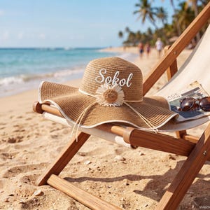 May include: A wide-brimmed straw hat with the name "Sokol" embroidered on the crown, adorned with a decorative flower, rests on a beach chair. Sunglasses and a magazine are also on the chair. The scene is set on a sandy beach with the ocean in the background.