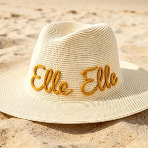 May include: A cream-coloured fedora-style sun hat with a wide brim. The hat features the name "Elle Elle" embroidered in a mustard-yellow, rope-like font across the front. The hat is resting on a sandy surface.